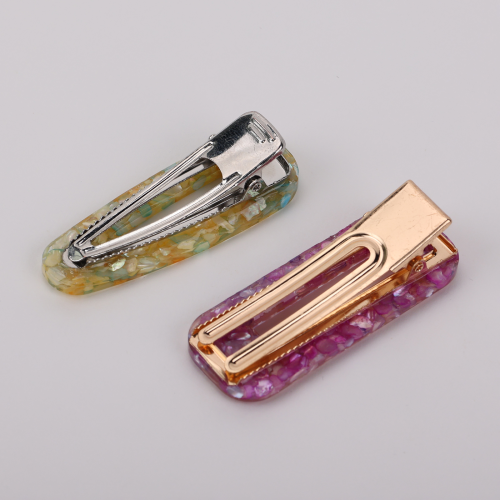 A pair of glossy resin snap hair clips in a pastel shell-inlay style, positioned as chic fashion hair accessories. One clip blends soft yellow, mint, and iridescent flecks with a silver-tone clasp, while the other features pink and lavender marbling with a warm gold-tone metal insert. Their rounded rectangular shape and luminous mosaic finish give them a feminine, polished feel suited to buyers drawn to playful elegance and boutique styling.
