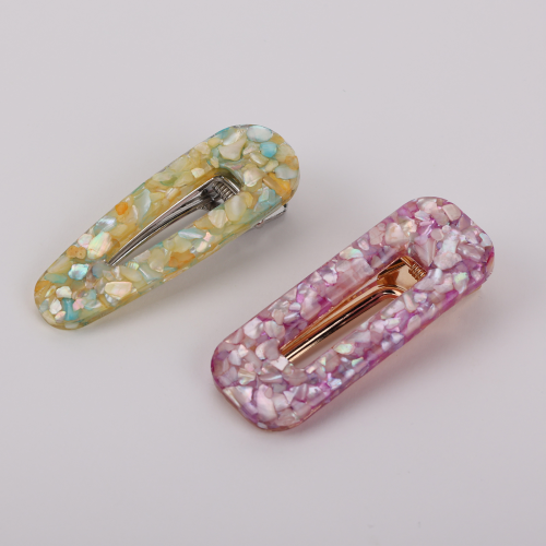A pair of glossy resin snap hair clips in a pastel shell-inlay style, positioned as chic fashion hair accessories. One clip blends soft yellow, mint, and iridescent flecks with a silver-tone clasp, while the other features pink and lavender marbling with a warm gold-tone metal insert. Their rounded rectangular shape and luminous mosaic finish give them a feminine, polished feel suited to buyers drawn to playful elegance and boutique styling.