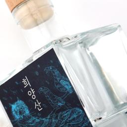 A clear square glass spirits bottle with a natural wooden cap, presented in a clean, minimalist premium style. The label is a striking dark panel with vivid blue line-art illustration and vertical Korean lettering, giving it an artisanal, contemporary edge. Its compact proportions, transparent glass, and restrained finish suggest a craft liquor or specialty soju aimed at design-conscious buyers. The overall mood is refined, modern, and gift-worthy.