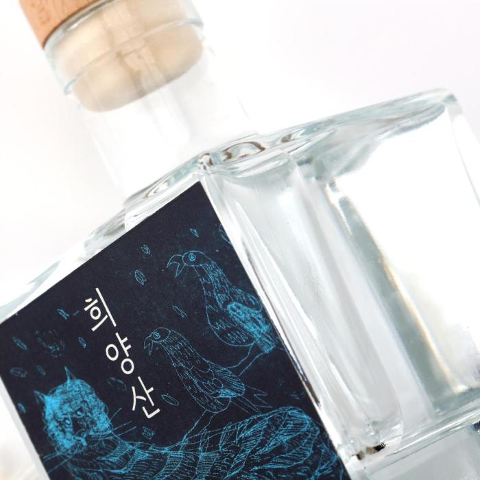 A clear square glass spirits bottle with a natural wooden cap, presented in a clean, minimalist premium style. The label is a striking dark panel with vivid blue line-art illustration and vertical Korean lettering, giving it an artisanal, contemporary edge. Its compact proportions, transparent glass, and restrained finish suggest a craft liquor or specialty soju aimed at design-conscious buyers. The overall mood is refined, modern, and gift-worthy.