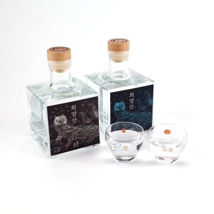 A clear square glass spirits bottle with a natural wooden cap, presented in a clean, minimalist premium style. The label is a striking dark panel with vivid blue line-art illustration and vertical Korean lettering, giving it an artisanal, contemporary edge. Its compact proportions, transparent glass, and restrained finish suggest a craft liquor or specialty soju aimed at design-conscious buyers. The overall mood is refined, modern, and gift-worthy.