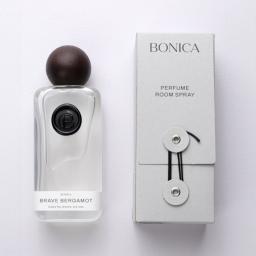 A refined perfume room spray from BONICA is presented as a clean, modern home fragrance product with a minimalist luxury feel. The bottle is a clear, softly rounded rectangular glass form topped with a smooth dark spherical cap, giving it a sculptural, artisanal look, while the black circular emblem on the front adds a subtle branded accent. The paired packaging box is light gray with understated typography, reading