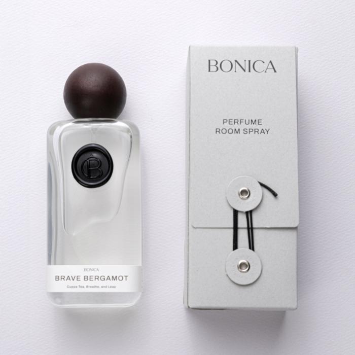 A refined perfume room spray from BONICA is presented as a clean, modern home fragrance product with a minimalist luxury feel. The bottle is a clear, softly rounded rectangular glass form topped with a smooth dark spherical cap, giving it a sculptural, artisanal look, while the black circular emblem on the front adds a subtle branded accent. The paired packaging box is light gray with understated typography, reading