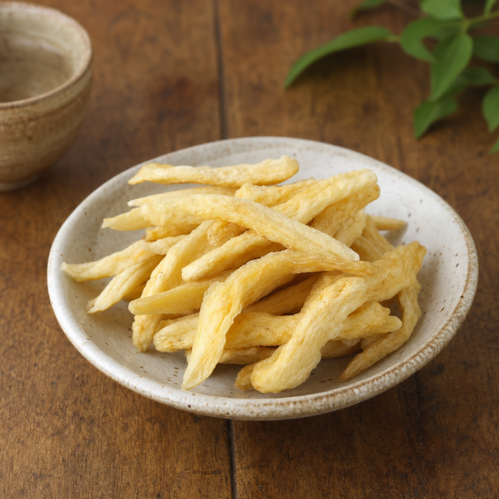 A rustic bowl of golden dried root snack pieces, likely a traditional pantry or tea-time delicacy, presented in a simple handcrafted ceramic dish. The strips have an irregular, naturally fibrous shape with a pale yellow tone and a lightly crisped, matte surface that suggests a chewy crunch. Set on a warm wooden table with understated background decor, the scene feels artisanal, wholesome, and quietly premium for buyers who appreciate natural snacks and traditional ingredients.